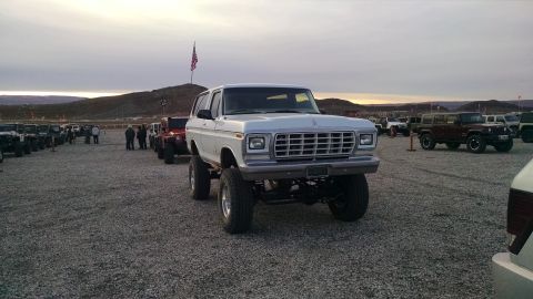 1979 Ford Bronco - A lifted white 1979 Ford Bronco is parked in a gravel lot with other off-road vehicles under a cloudy sky. An American flag flies on a pole…