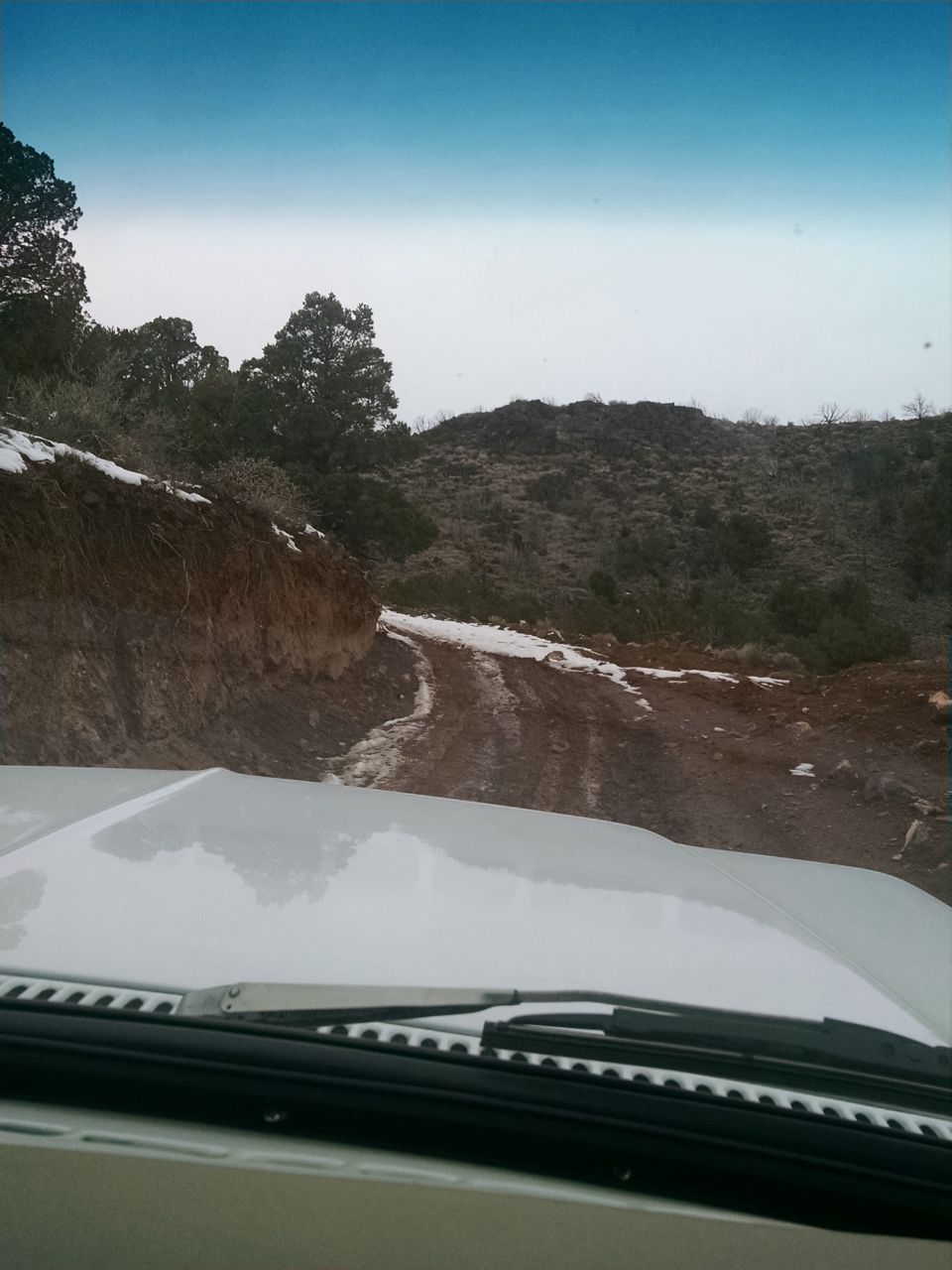 White vehicle driving on a muddy and snowy dirt road with trees and hills.