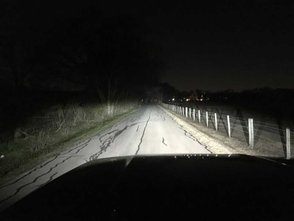 Vehicle's perspective driving on a dark, cracked road at night, with a fence and distant lights.