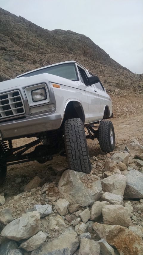 1979 Ford Bronco - A lifted white 1979 Ford Bronco is parked on a rocky, off-road terrain with a mountainous backdrop. The vehicle's front end, large tires, a…