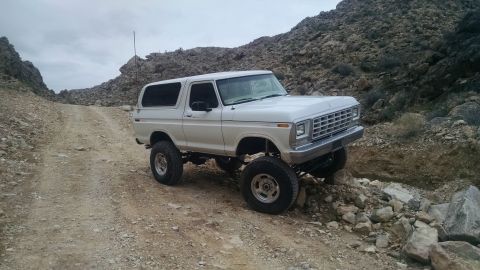 1979 Ford Bronco - A lifted white 1979 Ford Bronco is shown navigating a rocky, off-road trail under an overcast sky. The vehicle is positioned at an angle, s…