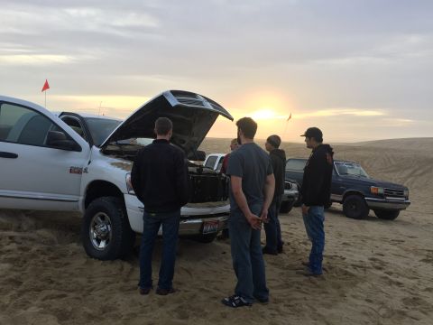 1989 Ford Bronco - Several people gather around a white pickup truck with its hood open as the sun sets over sand dunes.