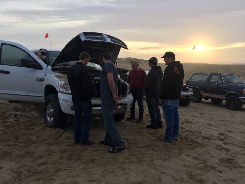 1989 Ford Bronco - A group of people inspect a white pickup truck with its hood open, while a dark blue SUV sits in the background on a sandy terrain.