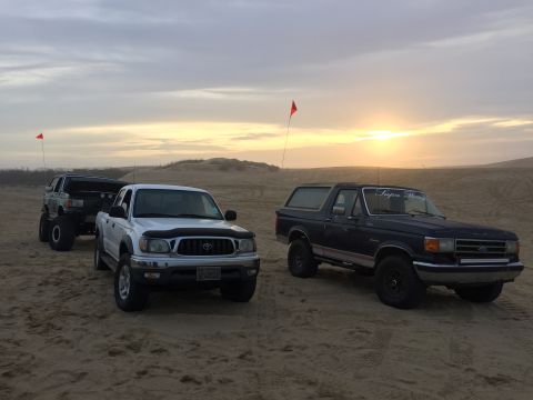1989 Ford Bronco - A dark blue 1989 Ford Bronco is parked on a sandy dune with a sunset in the background, alongside two other vehicles.
