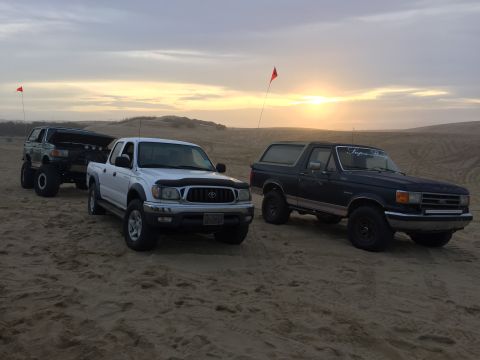 1989 Ford Bronco - Three trucks, including a 1989 Ford Bronco, parked on sand dunes during sunset.