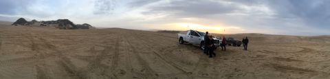 1989 Ford Bronco - Several vehicles are parked on sandy terrain with tire tracks visible, under a dramatic sky at sunset.