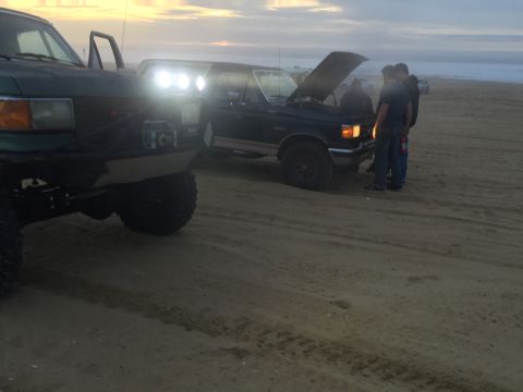 1989 Ford Bronco - Two SUVs parked on a sandy beach as the sun sets, one with its hood up.