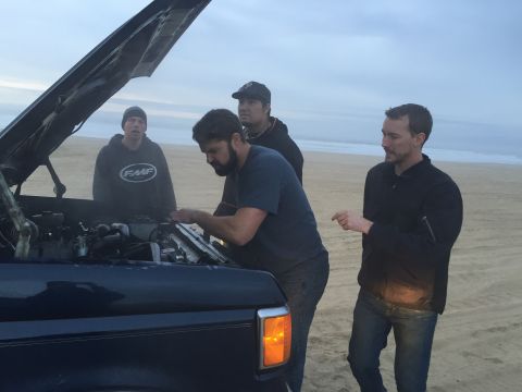 1989 Ford Bronco - A group of men inspect the engine of a vehicle on a beach under an overcast sky.
