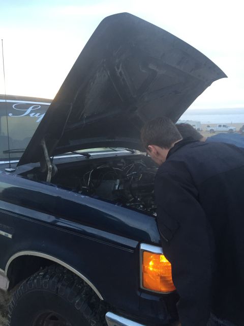 1989 Ford Bronco - Two people examine the engine of a dark blue Ford truck on a beach.