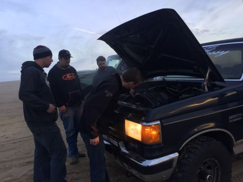 1989 Ford Bronco - A group of people inspect the engine of a dark blue 1989 Ford Bronco on a sandy terrain.