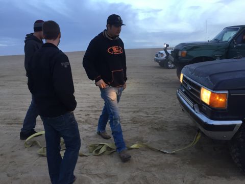 1989 Ford Bronco - A dark blue Ford F-Series truck is parked on sand with a tow strap connected to its front bumper.