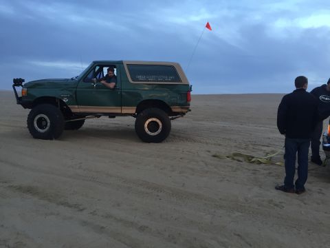 1989 Ford Bronco - A modified Ford Bronco sits on the sand, ready for off-road adventure under a cloudy sky.