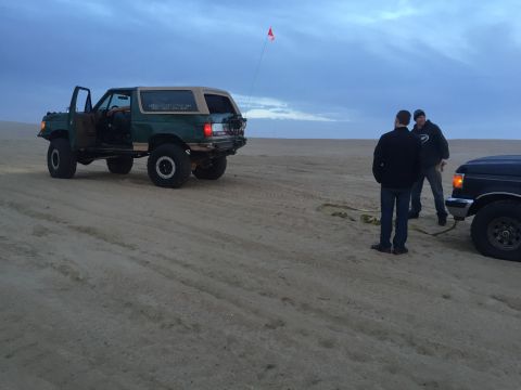 1989 Ford Bronco - Two men stand on a sandy beach next to a green Ford Bronco, with another vehicle partially visible in the foreground.