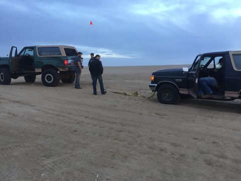 1989 Ford Bronco - Two Ford Broncos are on a sandy beach, one is being towed by the other. People are standing around.
