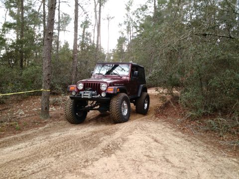 1992 Ford Bronco - This maroon Jeep Wrangler Rubicon navigates a sandy off-road trail through a wooded area.
