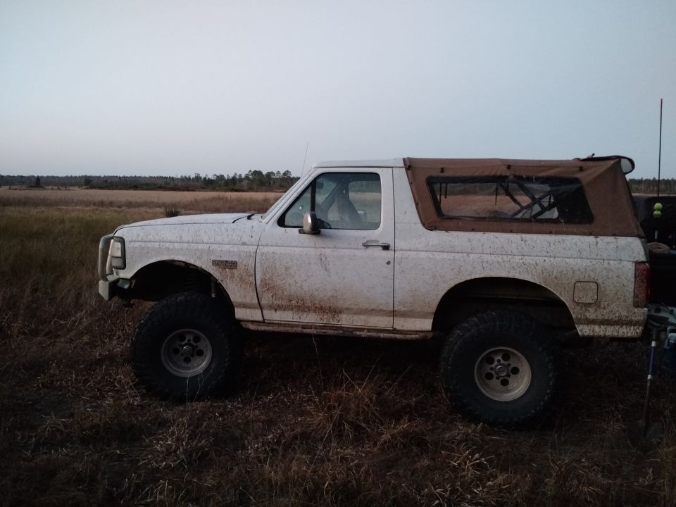 Side view of a white 1992 Ford Bronco with a brown soft top, covered in mud, parked in a grassy field