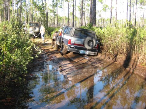 1994 Ford Bronco - Two Ford Broncos are stuck in a muddy forest trail, one partially submerged in water.