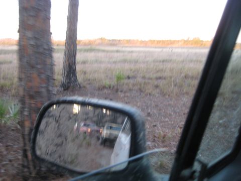 1994 Ford Bronco - Reflection of two SUVs on a dirt road in a side mirror, with trees and a field in the background.