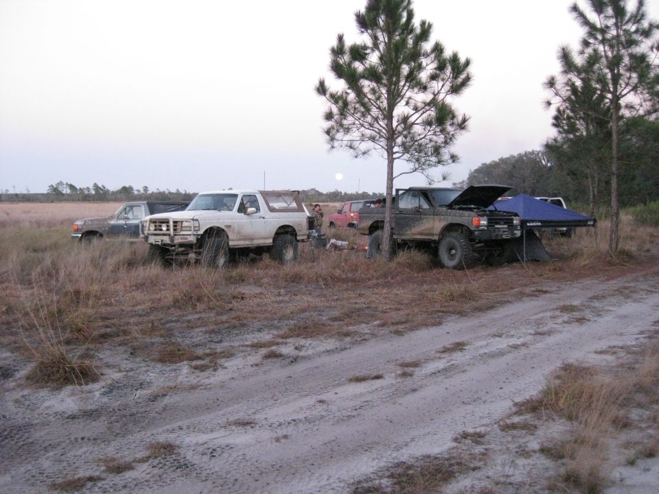 White 1992-1996 Ford Bronco parked in dry grass next to a black truck with hood open and a tent