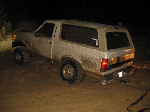 1994 Ford Bronco - This muddy Ford Bronco is parked on a dirt surface at night, with a tow strap connected to its rear.