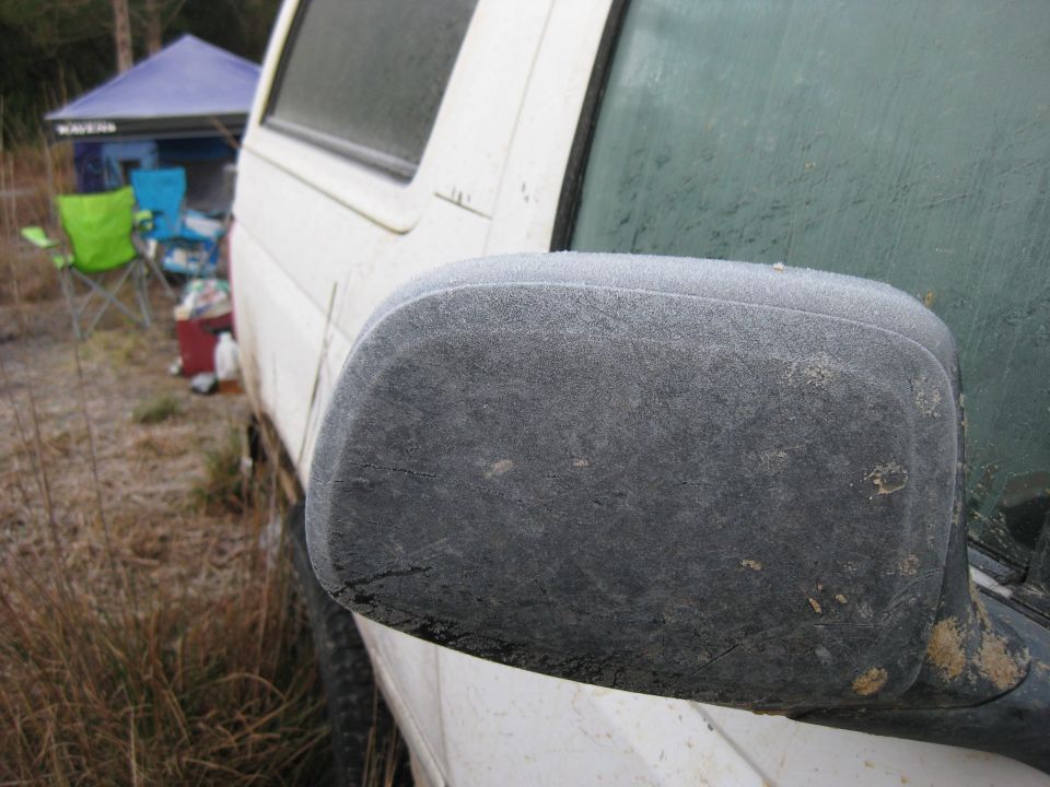 Frost covers the side mirror and part of the windshield of a white vehicle parked outdoors