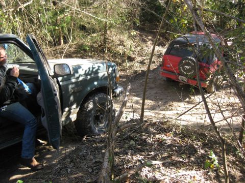 1994 Ford Bronco - Two SUVs navigate a muddy, wooded trail, with one vehicle stopped and its door open.