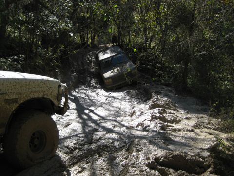 1994 Ford Bronco - Two off-road vehicles navigate a treacherous, muddy trail deep in a wooded area.