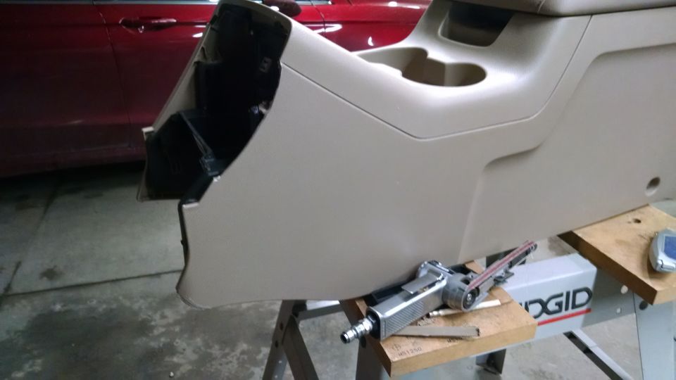 Center console removed from a vehicle, showing cup holders and a black interior cavity. A belt sander is on a workbench below
