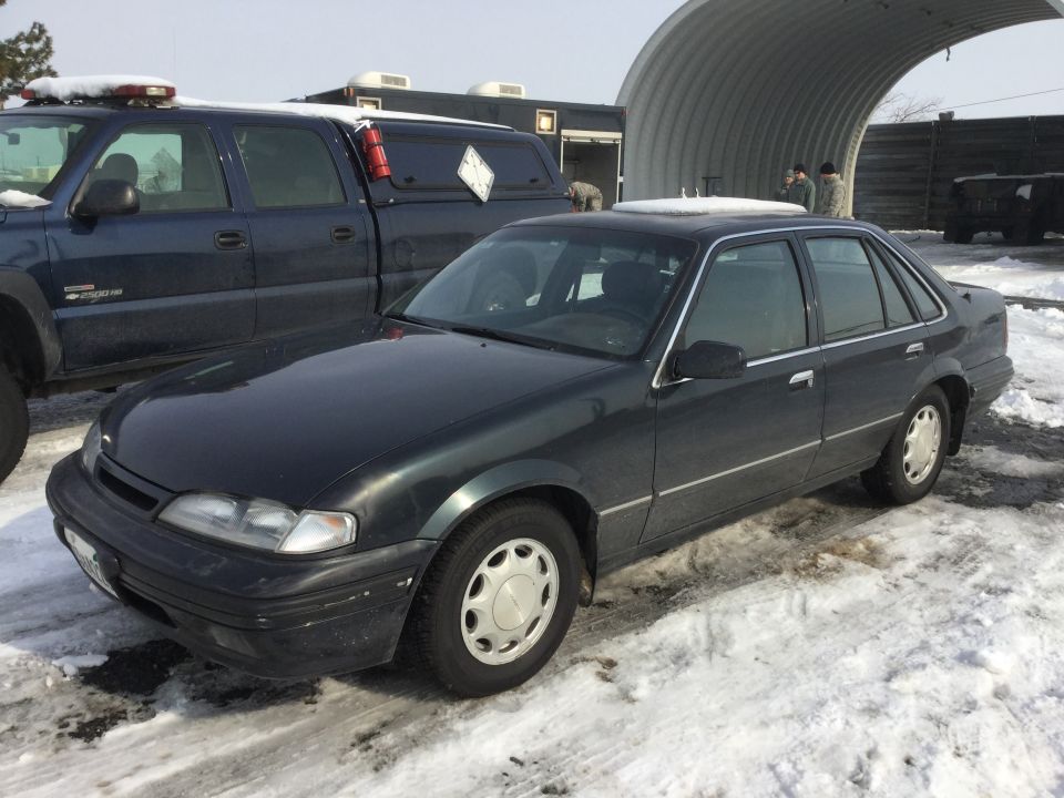 Dark sedan parked next to a blue pickup truck in a snowy lot. A large arched structure is in the background