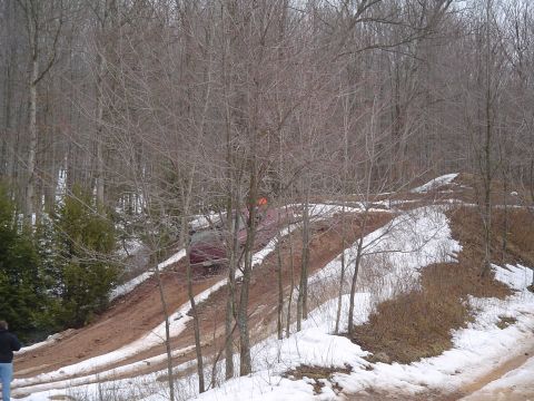 1993 Ford Bronco - A red SUV navigates a challenging muddy and snowy incline through a bare-branched forest.