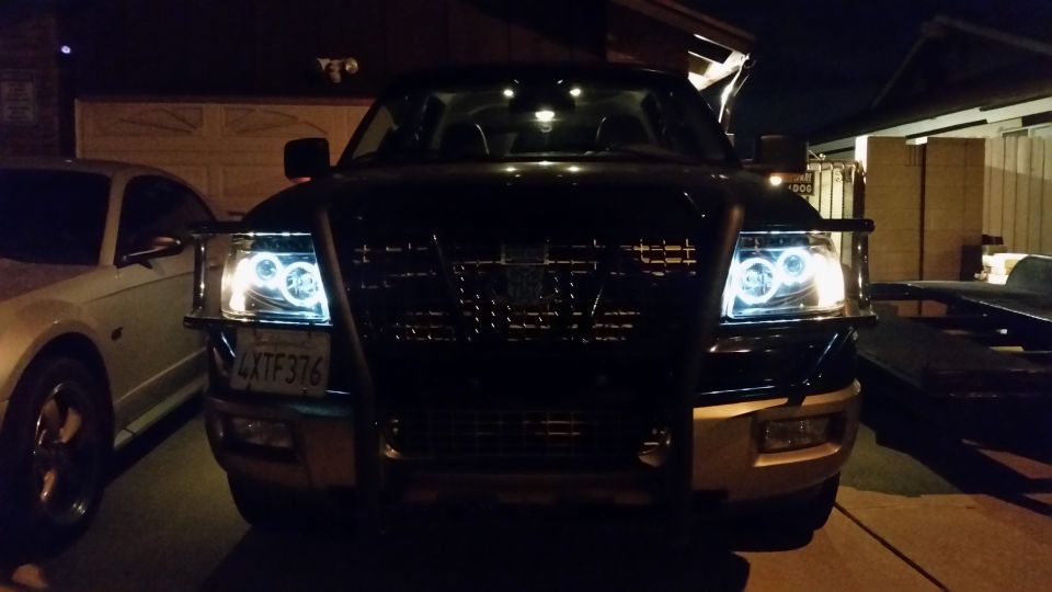 Front of a dark Ford F-150 with halo headlights and a brush guard. License plate reads 4XTF376. A silver car is parked to the left