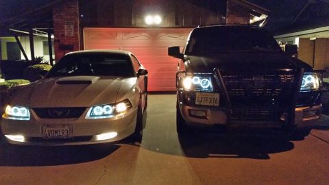 2003 Ford Crown Victoria - silver Ford Mustang coupe and a blacked-out SUV sit side by side in a residential driveway at night, both glowing with halo headlight rings.