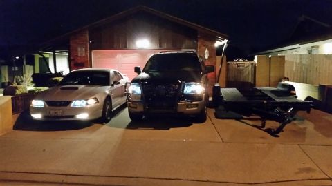 2003 Ford Crown Victoria - silver Ford Mustang and a dark SUV with a brush guard sit in a driveway at night beside an empty flatbed car trailer.