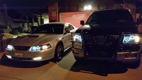 2003 Ford Crown Victoria - Close front view of a silver Ford Mustang and a black SUV with brush guard, both lit by bright halo-ring headlights in a night driveway.