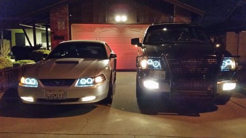 2003 Ford Crown Victoria - silver Ford Mustang and a dark SUV sit in a driveway at night, both with halo headlights and fog lights glowing.