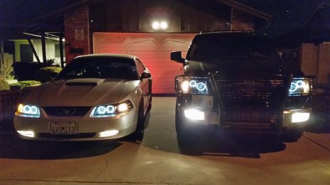 2003 Ford Crown Victoria - silver Ford Mustang and a pair of dark SUVs sit in a residential driveway at night, all lit by glowing halo headlights.