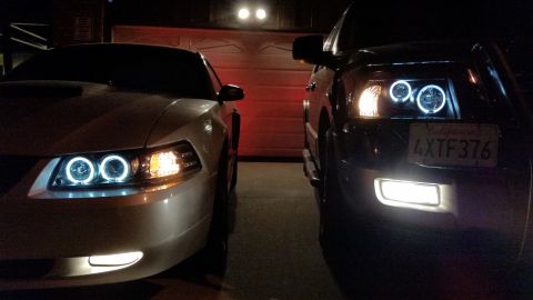 2003 Ford Crown Victoria - Tight close-up of the front corners of a silver car and a dark SUV at night, both fitted with bright halo-ring headlights.