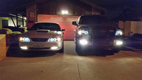 2003 Ford Crown Victoria - silver Ford Mustang and two dark SUVs sit in a driveway at night, all glowing with halo headlights and fog lamps.