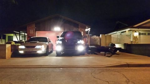 2003 Ford Crown Victoria - silver Ford Mustang and a dark SUV with bright fog lights sit in a driveway at night next to an empty flatbed trailer.