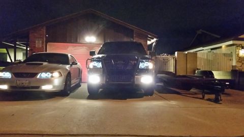 2003 Ford Crown Victoria - silver Ford Mustang and a black SUV with a brush guard and glowing halo headlights sit beside a flatbed trailer in a night driveway.