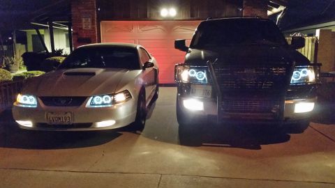 2003 Ford Crown Victoria - silver Ford Mustang and a blacked-out SUV with a brush guard glow with halo headlights and fog lamps in a residential driveway at night.