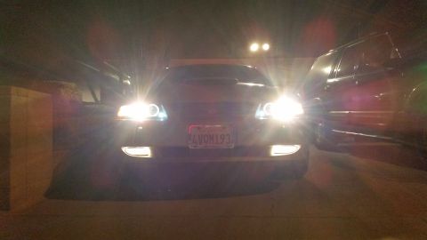 2002 Ford Mustang - A dark 2002 Ford Mustang is parked outside with its headlights illuminated.