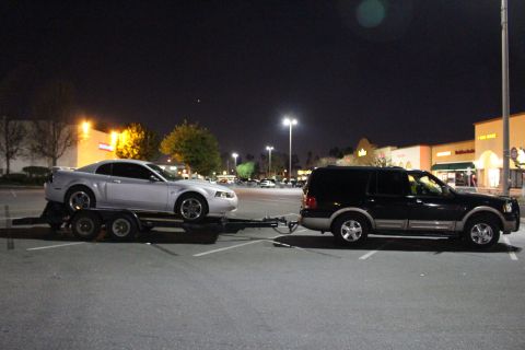 2002 Ford Mustang - A silver 2002 Ford Mustang coupe is transported on a trailer, towed by a black SUV in a parking lot at night.