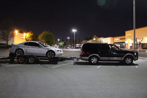 2002 Ford Mustang - A black Ford Expedition tows a silver Ford Mustang on a trailer at night in a shopping center parking lot.