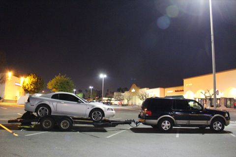 2002 Ford Mustang - A silver 2002 Ford Mustang is transported on a trailer towed by a dark SUV in a parking lot at night.