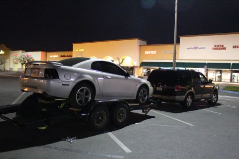 2002 Ford Mustang - A silver 2002 Ford Mustang is loaded onto a trailer, with a dark SUV towing it in a parking lot at night.