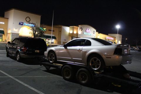2002 Ford Mustang - A white Ford Mustang is loaded onto a trailer, with a black SUV towing it in front of a strip mall at night.