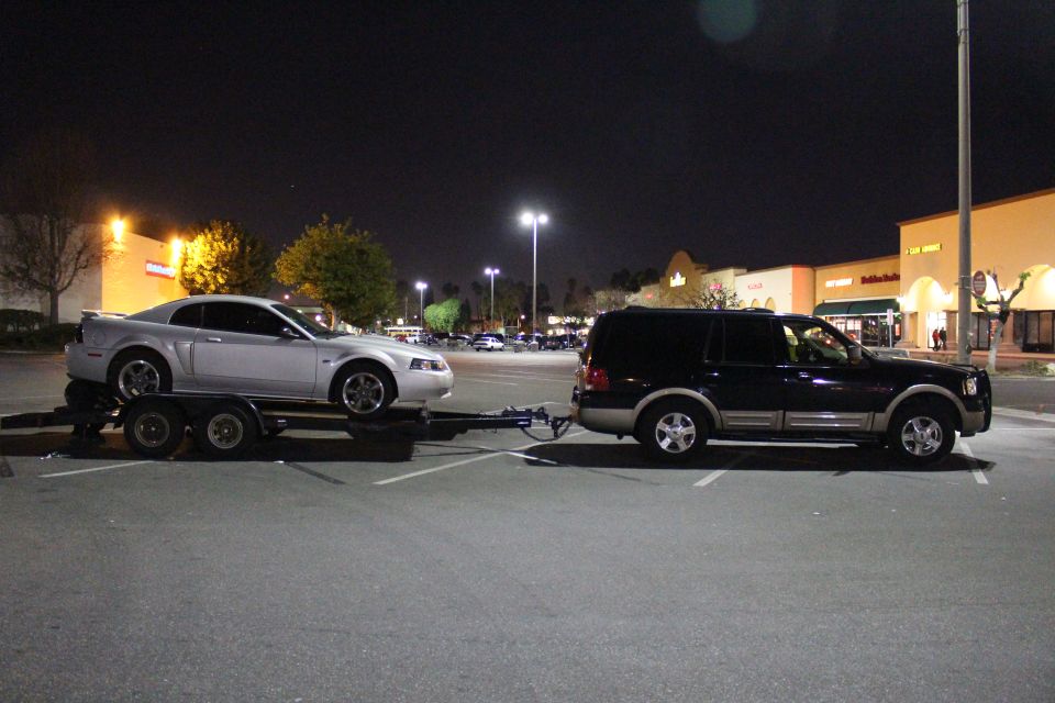 Dark blue Ford Expedition towing a silver Ford Mustang on a trailer at night in a parking lot