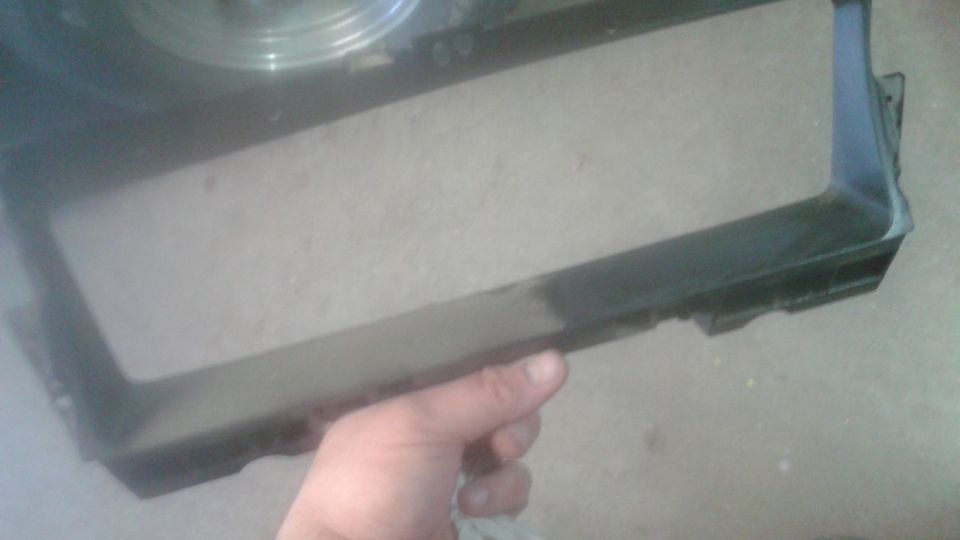 Hand holding a black plastic car grille frame. Part of a fan or radiator is visible above it