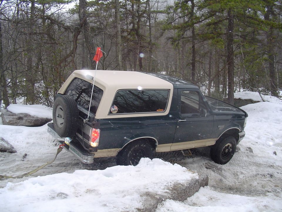 Dark green Ford Bronco with tan top in snow, spare tire on back, red flag on antenna